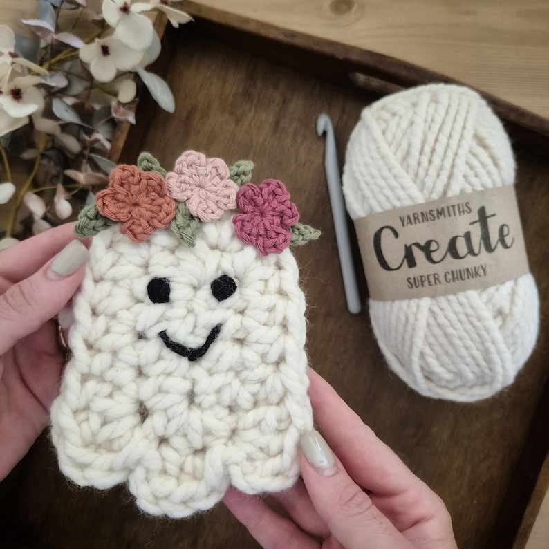 May include: A handmade, off-white crochet ghost with a smiling face and a floral crown. The ghost is held in front of a ball of cream-colored yarn with a label that reads "Yarnsmiths Create Super Chunky". A crochet hook is also visible.