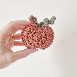 May include: A crocheted pumpkin with a green stem. The pumpkin is a light orange color and is made with a textured stitch.