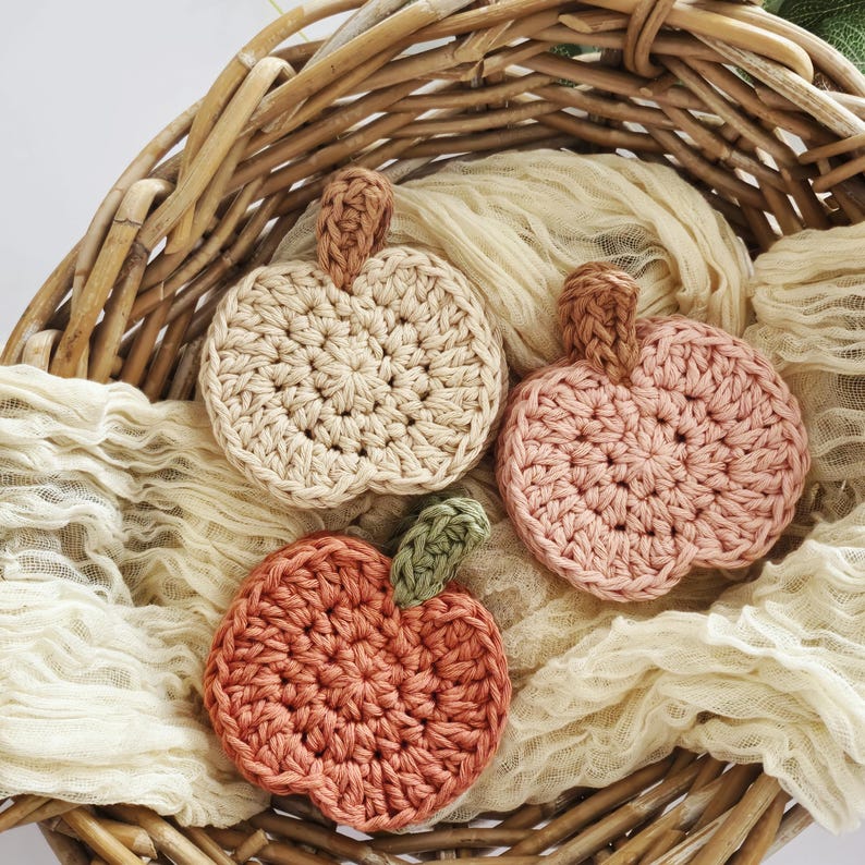 May include: Three handmade crochet pumpkin coasters in a wicker basket. The coasters are in shades of beige, pink, and orange, each with a brown or green stem. The pumpkins are arranged on a cream-colored fabric.
