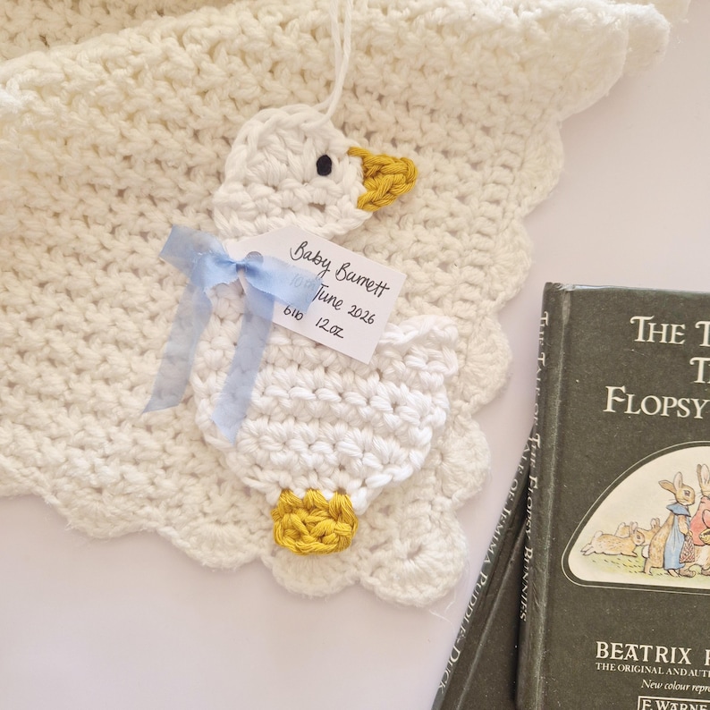 May include: A white crocheted blanket with a scalloped edge featuring a white crocheted duck applique with a yellow beak and feet. A light blue ribbon holds a white tag printed with Baby Barnett, June 2026, and 6lb 12oz. A book is visible nearby.