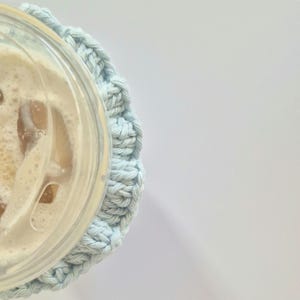 May include: A light blue crocheted cup sleeve wrapped around a clear plastic disposable cup containing an iced beverage with visible ice cubes and a clear plastic lid. The textured yarn sleeve features a repeating stitch pattern.