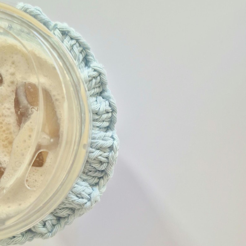 May include: A light blue crocheted cup sleeve wrapped around a clear plastic disposable cup containing an iced beverage with visible ice cubes and a clear plastic lid. The textured yarn sleeve features a repeating stitch pattern.