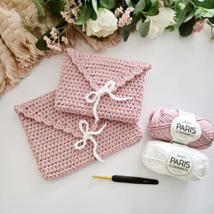 May include: Two pink crocheted envelope-style pouches with white ribbon closures. Two skeins of Drops Paris yarn, one pink and one white, and a crochet hook are also visible. The pouches have a scalloped edge.