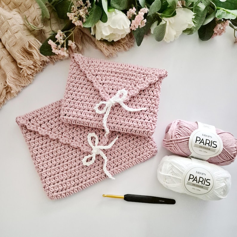 Puede incluir: Dos bolsos rosas de ganchillo con forma de sobre con cierres de cinta blanca. Tambi&eacute;n se ven dos madejas de hilo Drops Paris, una rosa y otra blanca, y un ganchillo. Los bolsos tienen un borde festoneado.