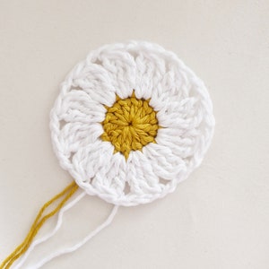 May include: A white crocheted daisy with a yellow center. The daisy is unfinished and has loose yarn ends.