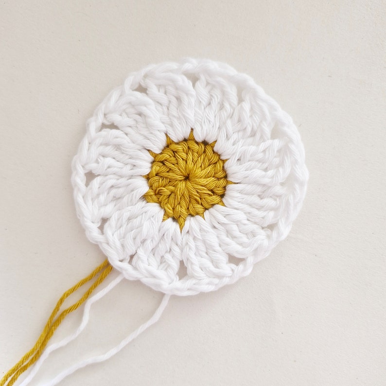 May include: A white crocheted daisy with a yellow center. The daisy is unfinished and has loose yarn ends.