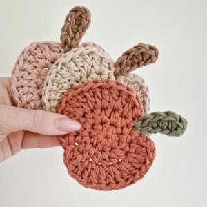 May include: A collection of handmade crochet coasters in the shape of pumpkins. The coasters are in shades of orange, pink, beige, and green. The crochet stitches are visible, and the coasters are held in a hand.