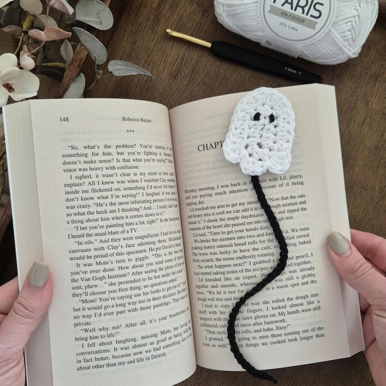 May include: A handmade white crochet ghost bookmark with black eyes and a black cord. The bookmark is placed in an open book with text on the pages. A ball of white yarn and a crochet hook are also visible.