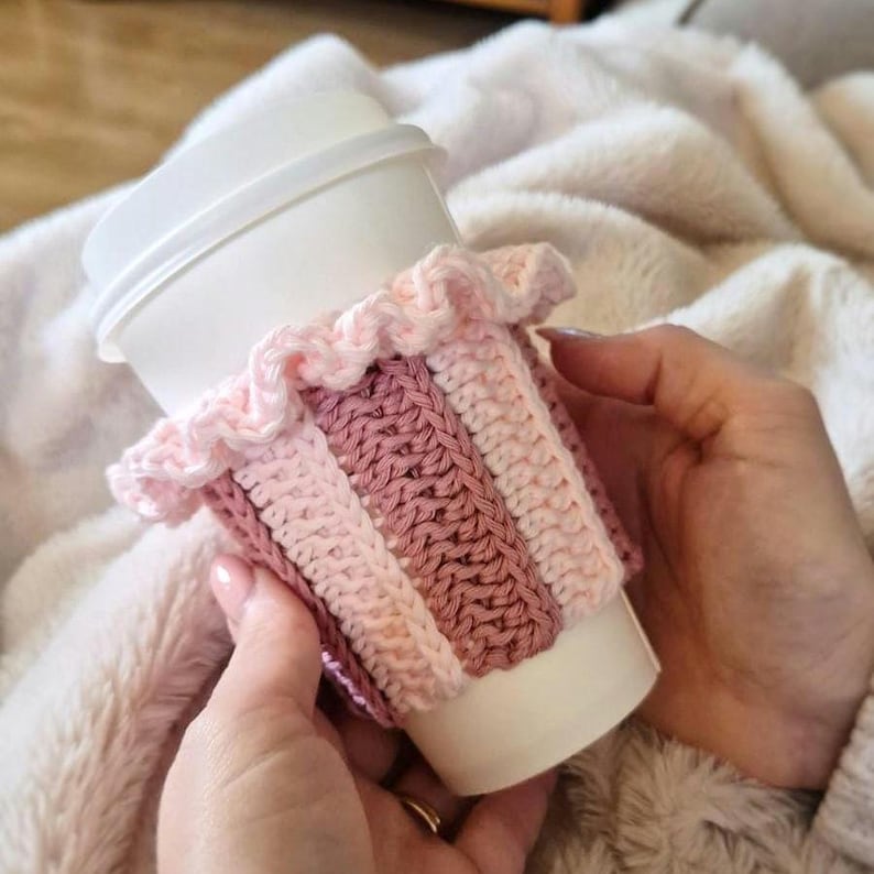 May include: A white disposable coffee cup featuring a crocheted sleeve with vertical stripes in shades of light pink, dusty rose, and cream. The sleeve includes a decorative ruffled edge at the top, providing a textured, layered appearance.