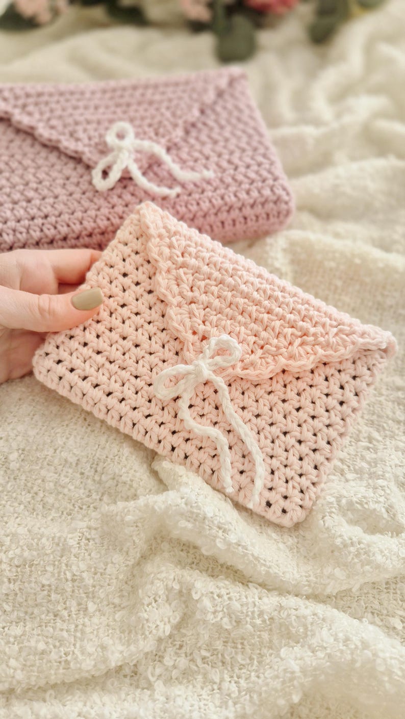 Puede incluir: Dos bolsos hechos a mano de ganchillo con forma de sobre en rosa suave. El bolso delantero est&aacute; enfocado, con un borde festoneado y un cierre de lazo de hilo blanco. El bolso de fondo tiene un dise&ntilde;o similar, ligeramente desenfocado.
