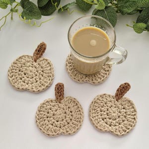 May include: Four handmade crochet pumpkin coasters in beige with brown stems. One coaster holds a clear glass mug filled with a light brown beverage. The coasters are arranged on a white surface, perfect for fall decor.