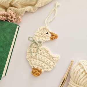 May include: A cream-colored crocheted goose ornament featuring a brown beak and feet, a sage green yarn bow tied around its neck, and a loop at the top for hanging. The goose is crafted from thick yarn with a visible textured stitch pattern.
