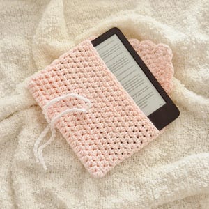 Puede incluir: Una funda de lector electr&oacute;nico de ganchillo rosa claro con cierre de cord&oacute;n blanco. La funda est&aacute; colocada sobre una manta texturizada de color crema. Un lector electr&oacute;nico es parcialmente visible dentro de la funda. Un peque&ntilde;o posavasos de ganchillo a juego est&aacute; al lado de la funda.