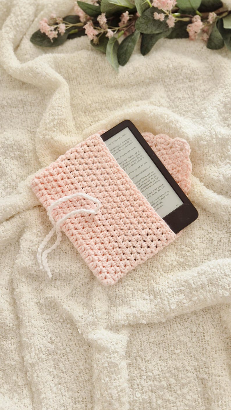 Puede incluir: Una funda de lector electr&oacute;nico de ganchillo rosa claro con cierre de cord&oacute;n blanco. La funda est&aacute; colocada sobre una manta texturizada de color crema. Un lector electr&oacute;nico es parcialmente visible dentro de la funda. Un peque&ntilde;o posavasos de ganchillo a juego est&aacute; al lado de la funda.