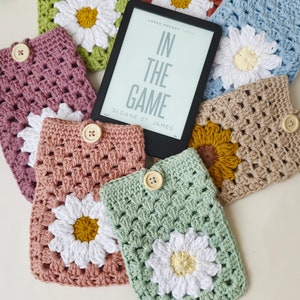 May include: A collection of crocheted daisy-themed book covers in various colors, including pink, green, beige, and blue. The covers feature a white daisy with a yellow center and are designed to fit an e-reader. The book covers are displayed on a white surface with a black e-reader displaying the title "In The Game" by Sloane St. James.