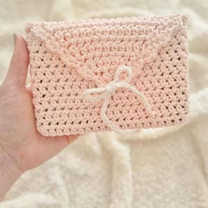 Puede incluir: Un bolso de mano de ganchillo rosa claro con un borde festoneado y un cierre de lazo blanco. El bolso hecho a mano es rectangular y parece estar hecho de hilo suave. El fondo es una tela texturizada de color crema.