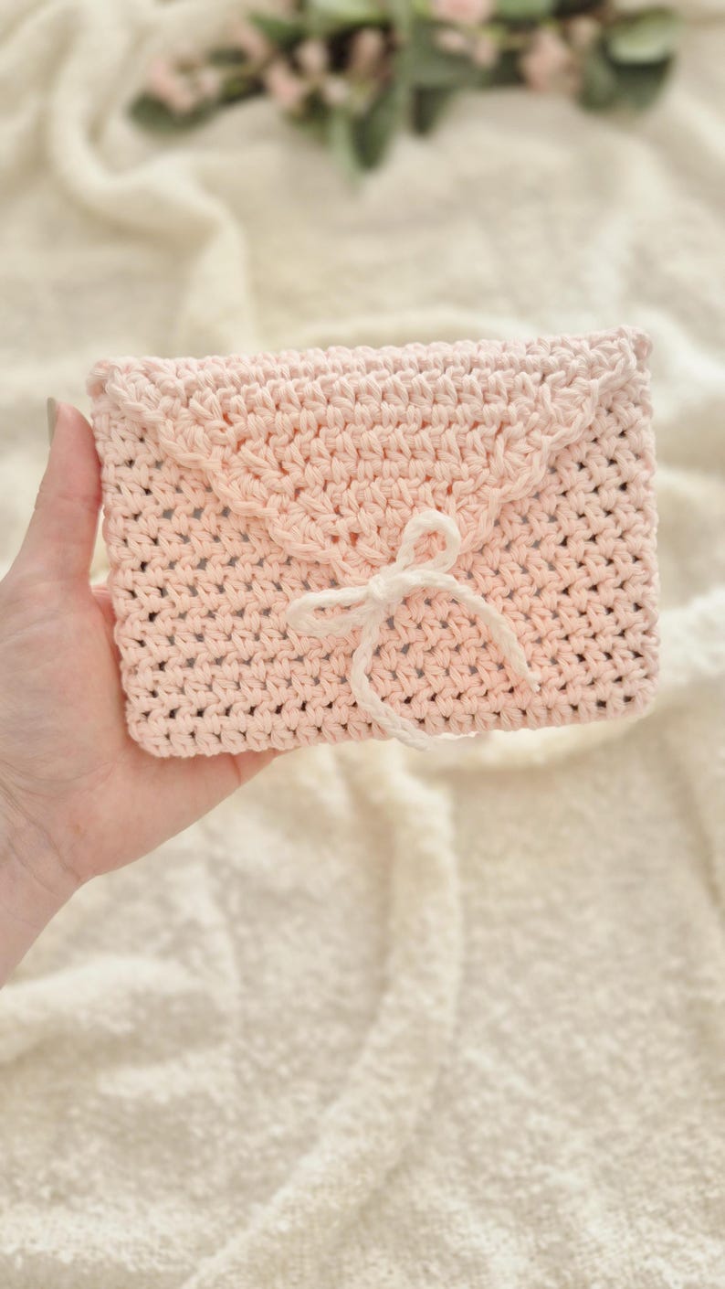 Puede incluir: Un bolso de mano de ganchillo rosa claro con un borde festoneado y un cierre de lazo blanco. El bolso hecho a mano es rectangular y parece estar hecho de hilo suave. El fondo es una tela texturizada de color crema.