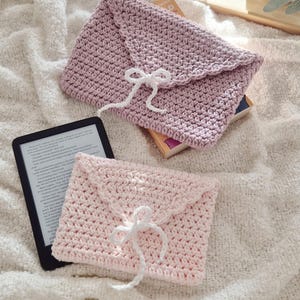 Puede incluir: Dos fundas hechas a mano de ganchillo con forma de sobre en rosa claro y malva, cada una con un cierre de lazo blanco. Un lector electr&oacute;nico es visible junto a la funda rosa. Las fundas est&aacute;n sobre una superficie blanca texturizada.