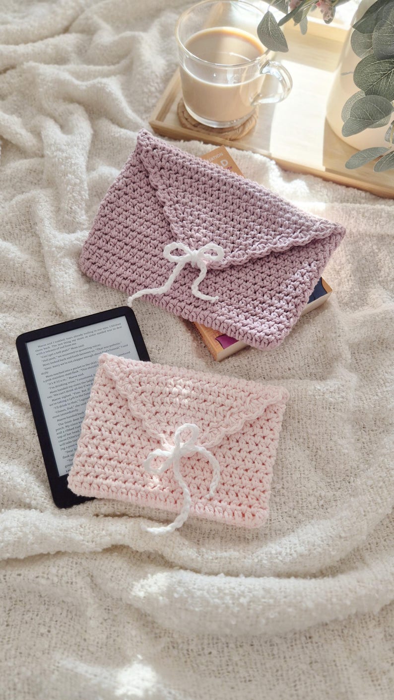 Puede incluir: Dos fundas hechas a mano de ganchillo con forma de sobre en rosa claro y malva, cada una con un cierre de lazo blanco. Un lector electr&oacute;nico es visible junto a la funda rosa. Las fundas est&aacute;n sobre una superficie blanca texturizada.