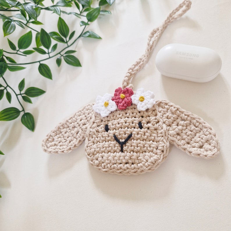 May include: A crocheted beige bunny with pink and white flowers on its head. The bunny is hanging from a string and has a black embroidered face.