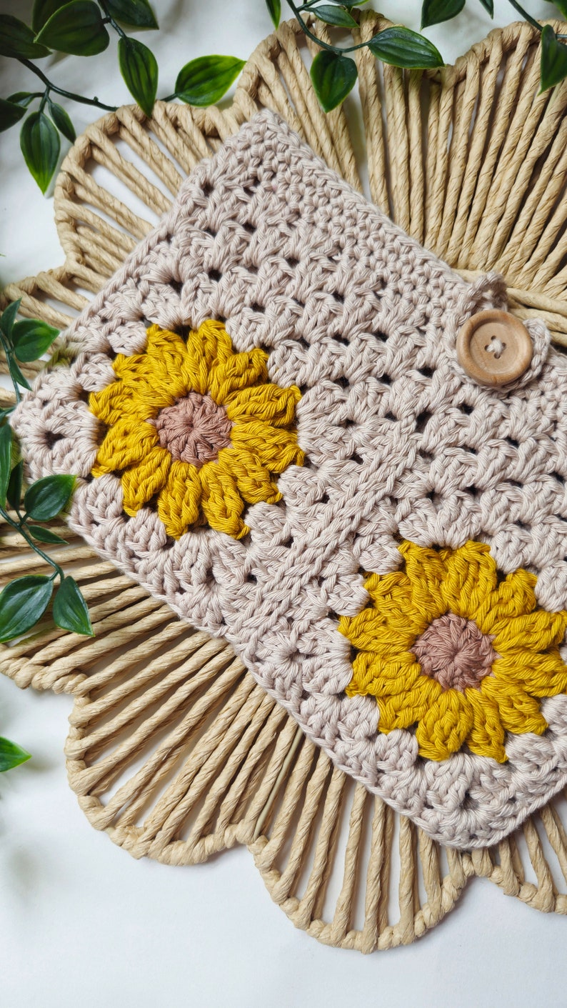 May include: A crocheted beige pouch with two yellow and brown crocheted sunflowers. The pouch has a wooden button closure.