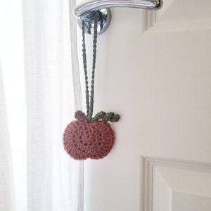 May include: A crocheted apple doorstop in shades of brown and green hanging from a white door with a silver door handle.