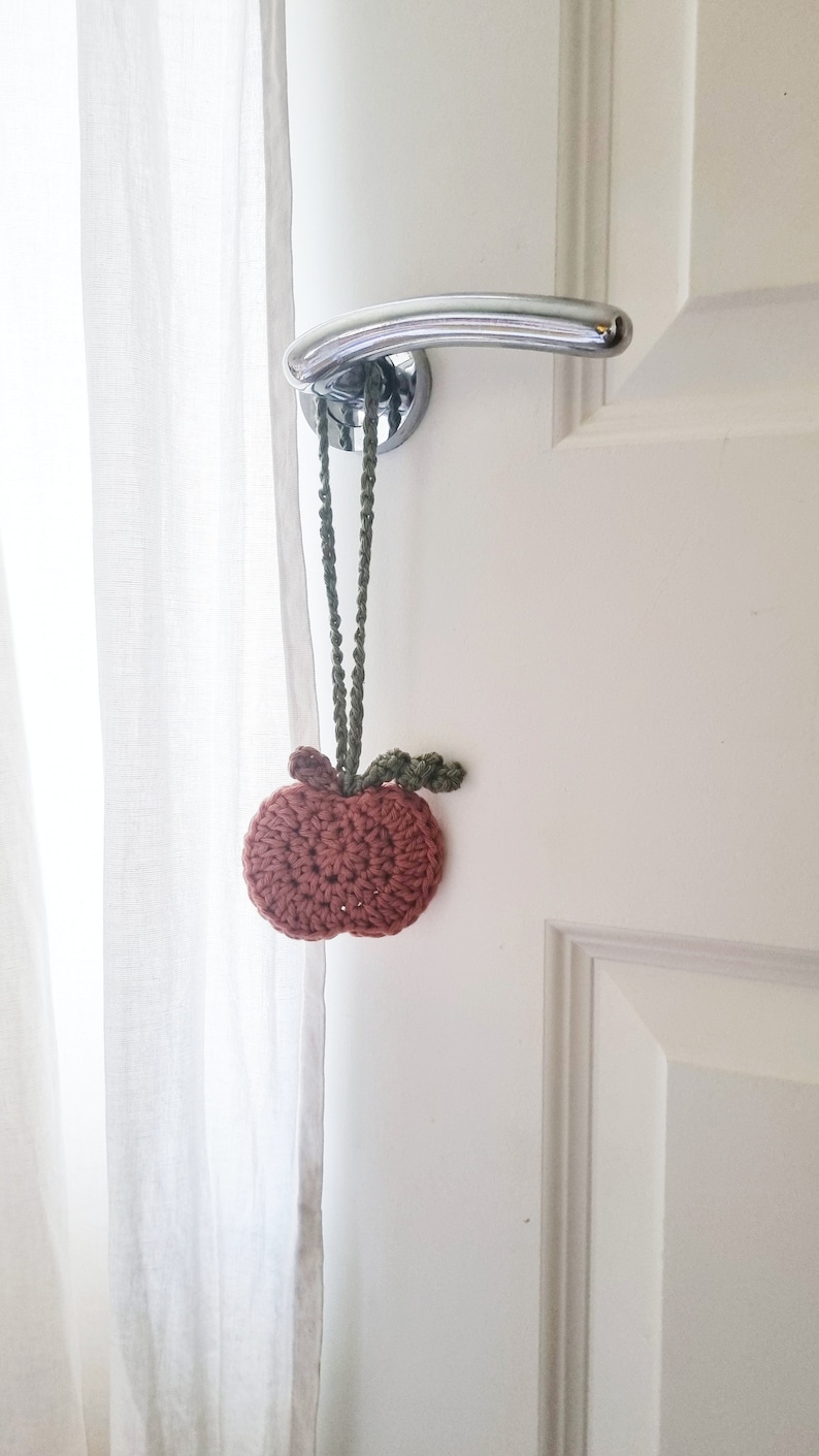 May include: A crocheted apple doorstop in shades of brown and green hanging from a white door with a silver door handle.