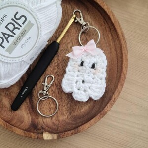 May include: A handmade white crochet ghost keychain with a pink bow, black eyes, and blush cheeks. A black and gold crochet hook, a silver keychain, and a ball of white yarn are also in the image. The yarn label reads "PARIS UNI COLOUR 100% Cotton".