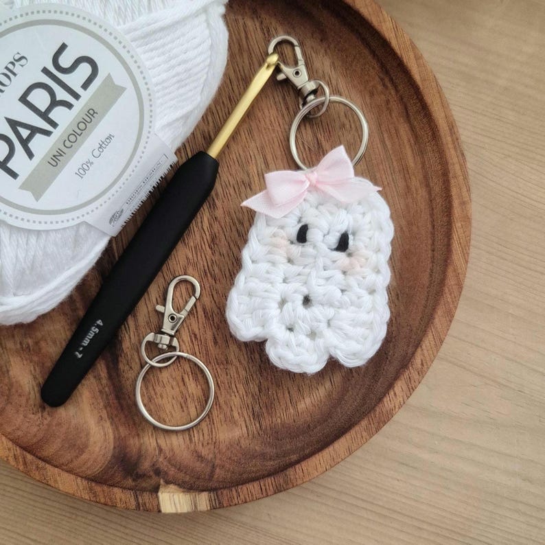 May include: A handmade white crochet ghost keychain with a pink bow, black eyes, and blush cheeks. A black and gold crochet hook, a silver keychain, and a ball of white yarn are also in the image. The yarn label reads "PARIS UNI COLOUR 100% Cotton".