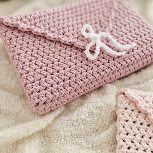 Puede incluir: Un bolso de mano de ganchillo rosa claro hecho a mano, estilo sobre, con solapa festoneada y cierre de lazo blanco. El bolso est&aacute; hecho de un hilo texturizado y se muestra sobre una superficie texturizada de color crema. Tambi&eacute;n se ve un art&iacute;culo m&aacute;s peque&ntilde;o a juego.