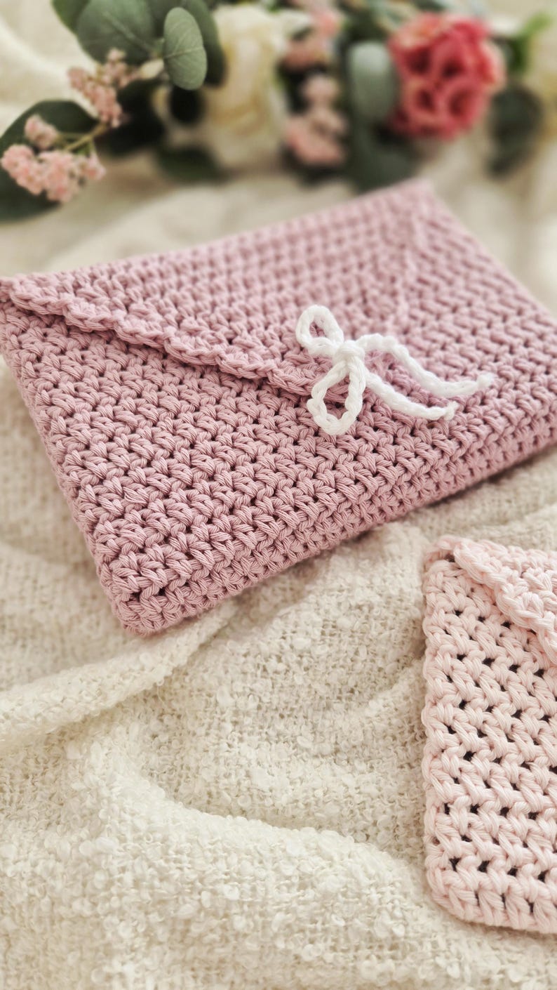 Puede incluir: Un bolso de mano de ganchillo rosa claro hecho a mano, estilo sobre, con solapa festoneada y cierre de lazo blanco. El bolso est&aacute; hecho de un hilo texturizado y se muestra sobre una superficie texturizada de color crema. Tambi&eacute;n se ve un art&iacute;culo m&aacute;s peque&ntilde;o a juego.