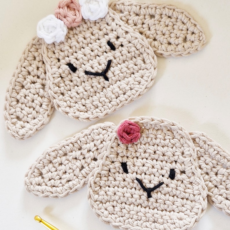 May include: Two crocheted beige lamb ornaments with embroidered faces. One lamb has three white and pink flowers on its head, the other has one pink flower.