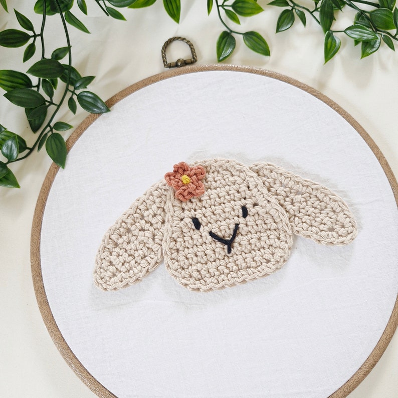 May include: A beige crocheted animal head with a pink flower on top, displayed on a white fabric background in a wooden embroidery hoop.