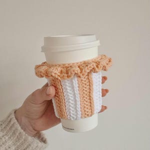 May include: A white disposable coffee cup featuring a removable, hand-crocheted sleeve. The sleeve is designed with vertical stripes in alternating peach and white yarn, finished with a decorative ruffled edge around the top rim.
