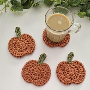 May include: Four handmade crochet pumpkin coasters in shades of orange and green. One coaster is under a glass mug filled with a light brown beverage. The coasters are a fall-themed home decor item.