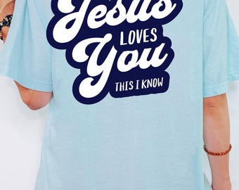 May include: Light blue t-shirt with the text "Jesus Loves You This I Know" in a white and navy blue design on the back. The shirt has short sleeves and a relaxed fit.