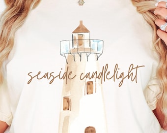 May include: Cream-coloured t-shirt featuring a watercolour illustration of a lighthouse with the words "seaside candlelight" in brown script. The lighthouse is beige and brown with a blue top. The shirt has short sleeves.