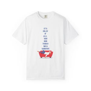 May include: A white short-sleeved t-shirt featuring a graphic of a person lying on a red sofa. Above the sofa, the text reads: IT'S OKAY IF ALL YOU DID TODAY WAS SURVIVE. The shirt has a crew neckline and a standard fit, presented on a plain background.