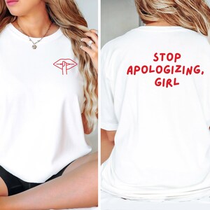 May include: White t-shirt with a red lip and finger graphic on the front. The back of the shirt has the text "STOP APOLOGIZING, GIRL" in red. The shirt is made of a soft material.