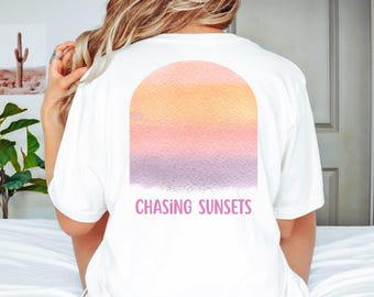 May include: White t-shirt with a watercolor design of a sunset in shades of orange, pink, and purple. The text "CHASING SUNSETS" is printed in pink below the design. The shirt is a casual style.