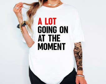 May include: White t-shirt with the text "A LOT GOING ON AT THE MOMENT" in black and red. The shirt is a casual, oversized style.