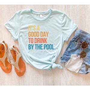 May include: A light blue t-shirt with the text "IT'S A GOOD DAY TO DRINK BY THE POOL" in orange, red, and blue. The shirt is paired with denim shorts, sunglasses, and orange sandals.