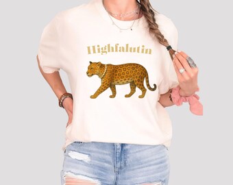 May include: Cream-coloured t-shirt featuring a leopard illustration with a pearl necklace and the word "Highfalutin" in gold lettering. The leopard has a spotted pattern in shades of brown and tan.