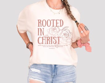 May include: Cream-colored t-shirt with the text "ROOTED IN CHRIST" in a rose-colored font, accompanied by rose illustrations. The shirt also features a Bible verse. The model is wearing blue jeans.