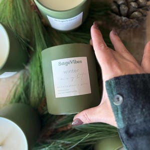 May include: Three green soy wax blend candles with white labels. The labels read "SageVibes" and "winter light - eucalyptus + pine" .