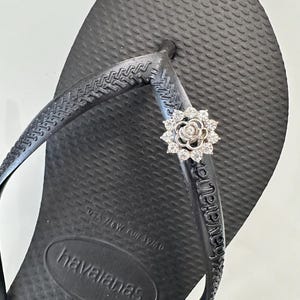 Pode incluir: Sand&aacute;lias Havaianas pretas com uma flor de strass na tira. As sand&aacute;lias t&ecirc;m o nome da marca "havaianas" impresso na sola.