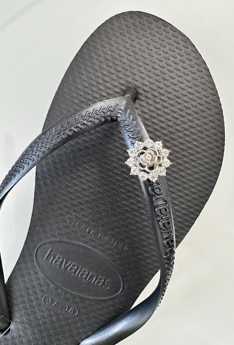 Pode incluir: Sand&aacute;lias Havaianas pretas com uma flor de strass na tira. As sand&aacute;lias t&ecirc;m o nome da marca "havaianas" impresso na sola.