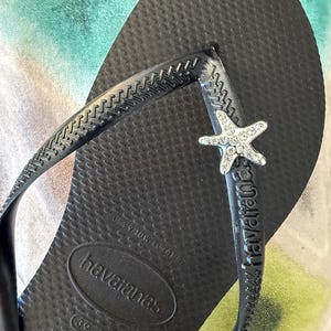 K&ouml;nnte beinhalten: Schwarze Havaianas-Zehensandale mit strukturierter Fu&szlig;sohle und Riemen. Der Riemen hat einen silbernen Seestern-Charm mit klaren Strasssteinen. Der Markenname ist auf Fu&szlig;bett und Riemen sichtbar.