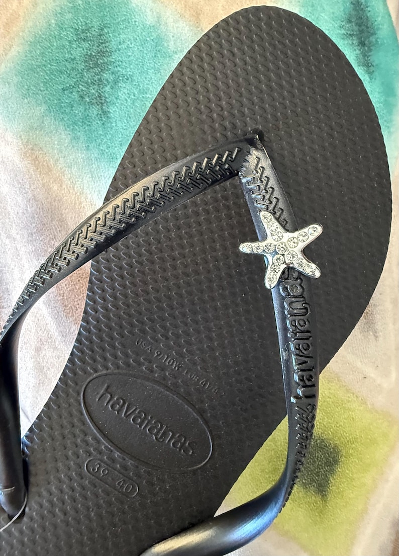 K&ouml;nnte beinhalten: Schwarze Havaianas-Zehensandale mit strukturierter Fu&szlig;sohle und Riemen. Der Riemen hat einen silbernen Seestern-Charm mit klaren Strasssteinen. Der Markenname ist auf Fu&szlig;bett und Riemen sichtbar.