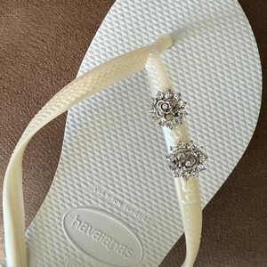 以下が含まれることがあります： 白のハワイアナスのビーチサンダル。ソールはテクスチャード加工で、クリーム色のストラップが付いています。ストラップには2つの銀色の花の装飾が施されています。サイズはEUR 41/42です。